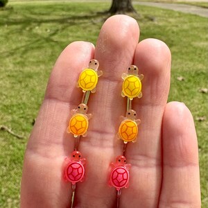 Puede incluir: Un juego de seis pinzas para el pelo de tortuga de colores. Las pinzas est&aacute;n hechas de pl&aacute;stico y tienen un acabado brillante. Las tortugas est&aacute;n dispuestas en un degradado de amarillo a naranja a rosa.