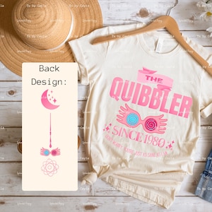 May include: Cream-coloured t-shirt with a pink graphic that reads "THE QUIBBLER SINCE 1980." The design includes a banner, glasses, and stars. The back of the shirt features a pink wand, glasses, a moon, stars, and an atom symbol.