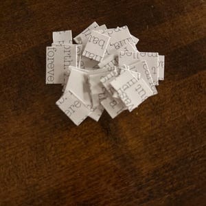 May include: A collection of small, white paper squares, each bearing printed text fragments such as "forever" and "birth." The squares are arranged on a dark wooden surface, creating a layered texture. The text appears to be from a book.