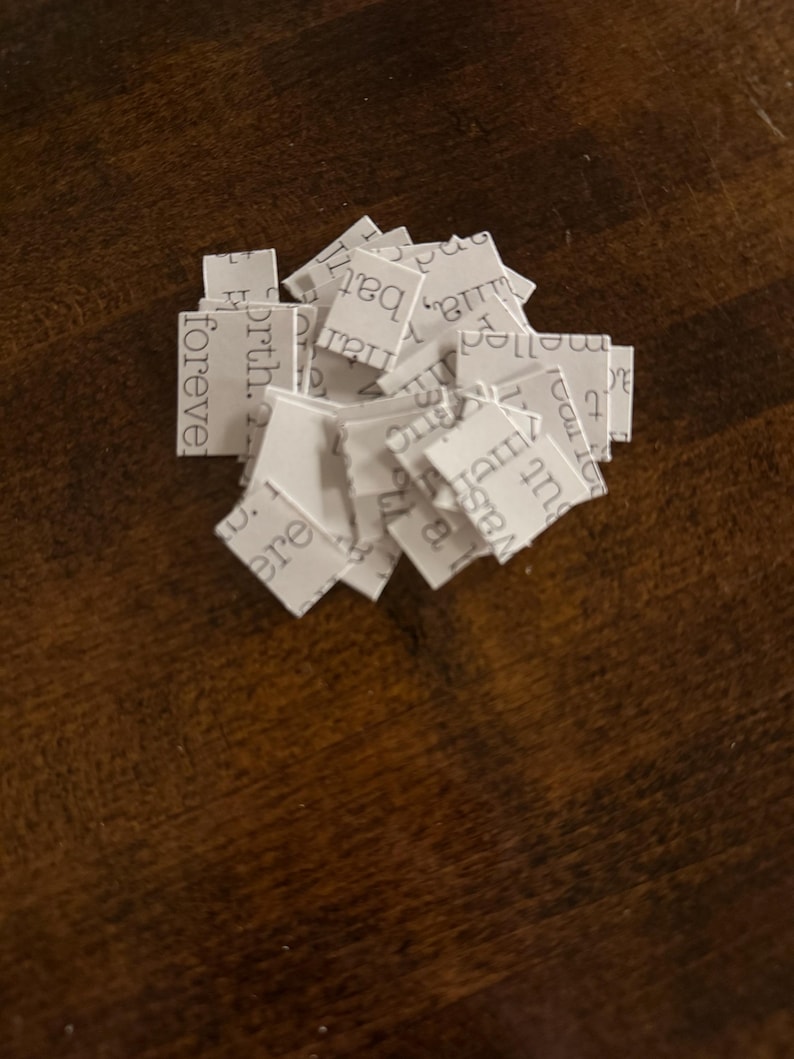May include: A collection of small, white paper squares, each bearing printed text fragments such as "forever" and "birth." The squares are arranged on a dark wooden surface, creating a layered texture. The text appears to be from a book.