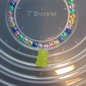 May include: A colourful beaded bracelet with a lime green gummy bear charm. The bracelet features clear, iridescent beads in various colours, including blue, pink, yellow, and green. The bracelet is approximately 18 cm in diameter.