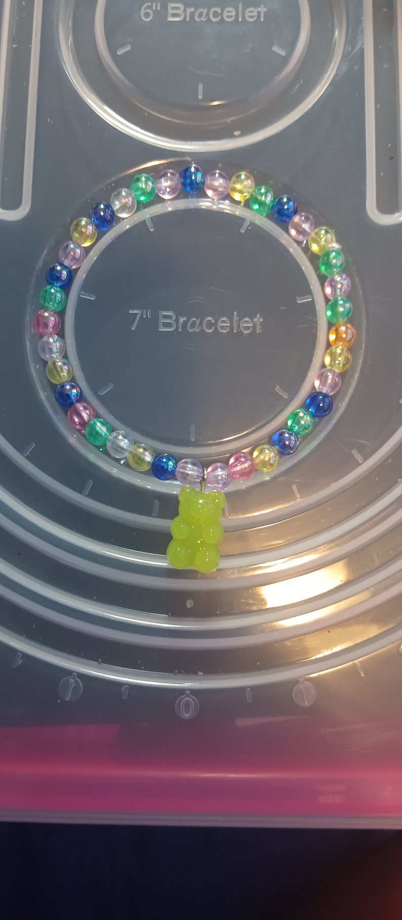 May include: A colourful beaded bracelet with a lime green gummy bear charm. The bracelet features clear, iridescent beads in various colours, including blue, pink, yellow, and green. The bracelet is approximately 18 cm in diameter.