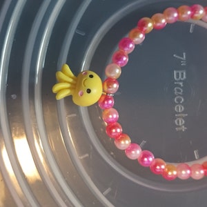 May include: A beaded bracelet with pink and orange beads and a yellow octopus charm. The bracelet is approximately 17.78 cm in diameter.