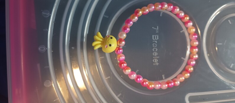 May include: A beaded bracelet with pink and orange beads and a yellow octopus charm. The bracelet is approximately 17.78 cm in diameter.