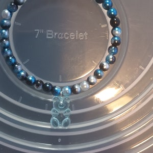 May include: A bracelet featuring blue, black, and silver beads, with a clear gummy bear charm. The background shows the text "7" Bracelet". The bracelet is approximately 17.78 cm in length.