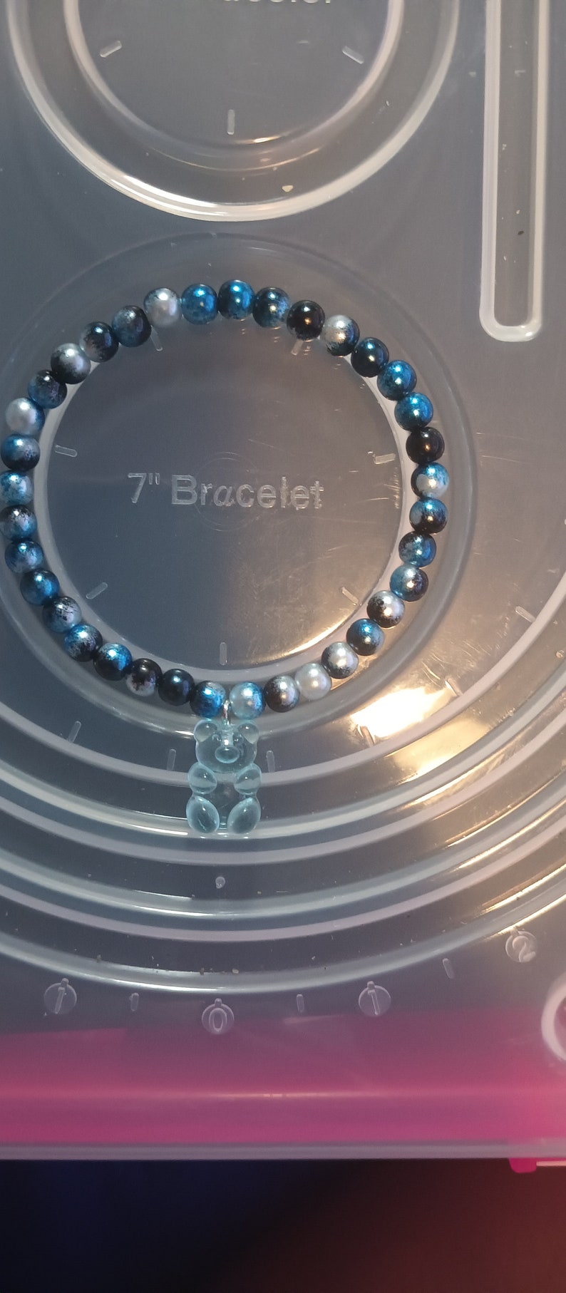 May include: A bracelet featuring blue, black, and silver beads, with a clear gummy bear charm. The background shows the text "7" Bracelet". The bracelet is approximately 17.78 cm in length.