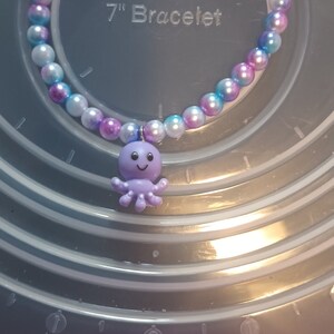 May include: A beaded bracelet with a purple octopus charm. The bracelet is made of round, iridescent beads in shades of white, blue, and purple. The text "7" Bracelet" is visible.