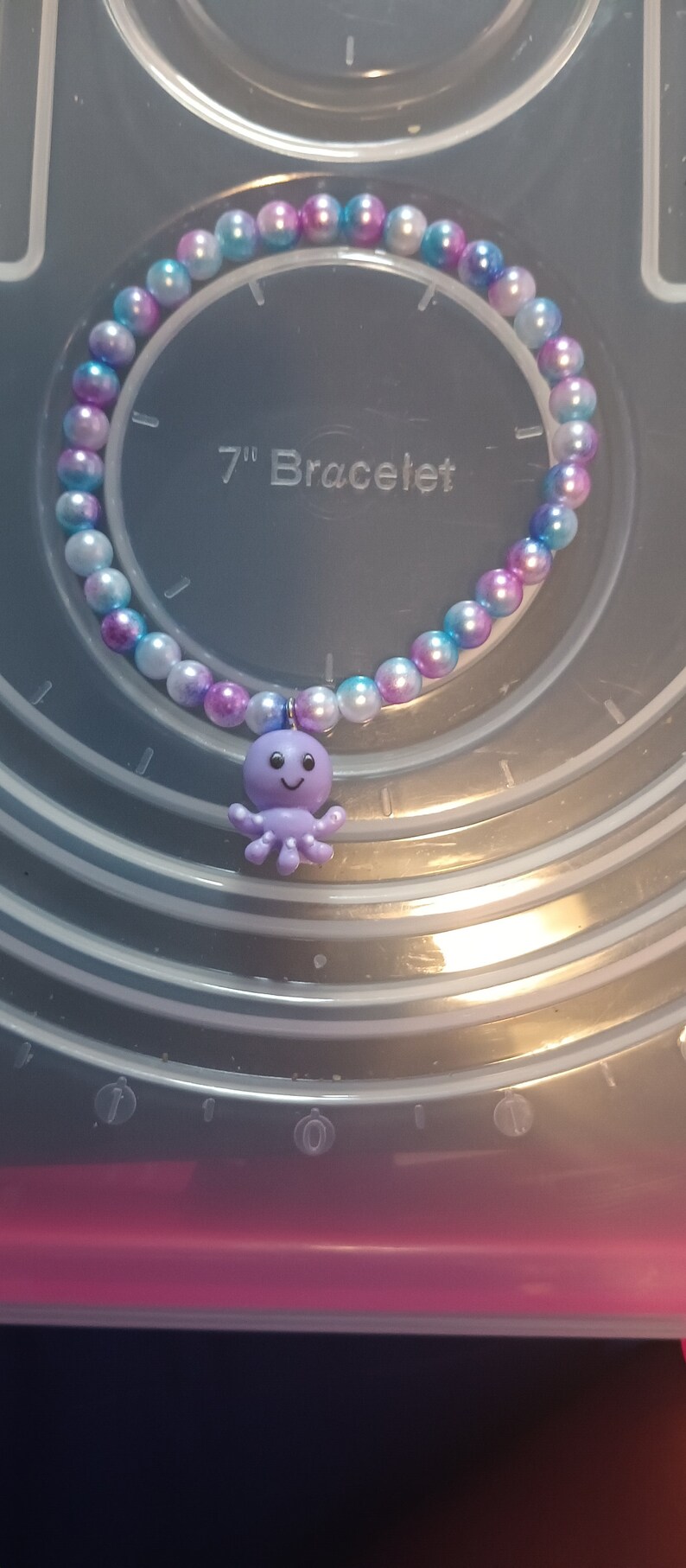 May include: A beaded bracelet with a purple octopus charm. The bracelet is made of round, iridescent beads in shades of white, blue, and purple. The text "7" Bracelet" is visible.