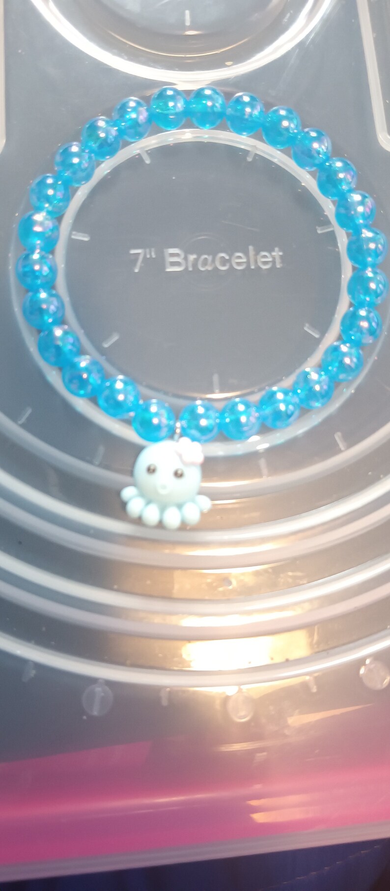 May include: A blue beaded bracelet with an octopus charm. The bracelet is approximately 17.78 cm in length. The charm is light blue with a pink bow.