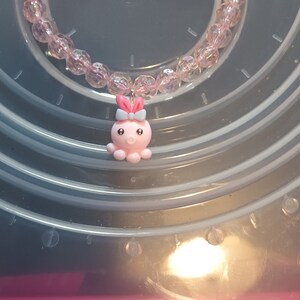 May include: A pink beaded bracelet, approximately 17.8 cm in circumference, with a charm of a pink octopus and a blue bow. The beads are faceted and translucent. The bracelet is displayed on a clear plastic surface.