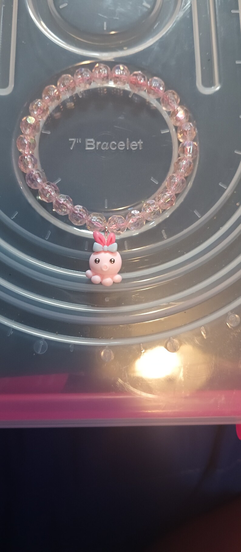 May include: A pink beaded bracelet, approximately 17.8 cm in circumference, with a charm of a pink octopus and a blue bow. The beads are faceted and translucent. The bracelet is displayed on a clear plastic surface.