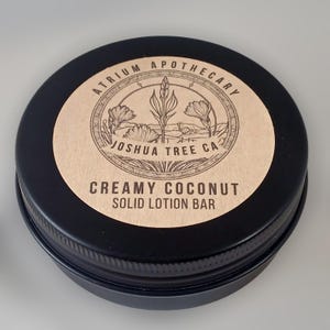 May include: A black tin container with a brown label that reads "Atrium Apothecary Joshua Tree CA Creamy Coconut Solid Lotion Bar".