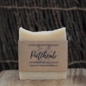 May include: A bar of white goat milk soap with a brown paper label that reads "Atrium Apothecary Patchouli Artisan Goat Milk Soap Handcrafted in 29 Palms CA."
