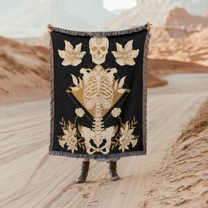 Könnte beinhalten: Eine schwarze gewebte Wandbehang mit einem goldenen Skelett-Design und floralen Akzenten. Das Skelett ist mit einem Schädel oben und floralen Elementen dargestellt, die es umgeben.