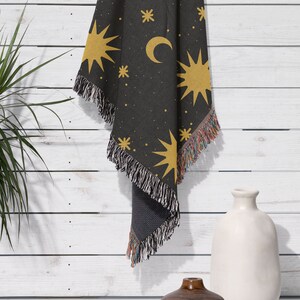 K&ouml;nnte beinhalten: Eine schwarze gewebte Wolldecke mit einem Muster aus gelben Sternen und Mondsicheln. Die Decke h&auml;ngt an einer wei&szlig;en Holzwand.