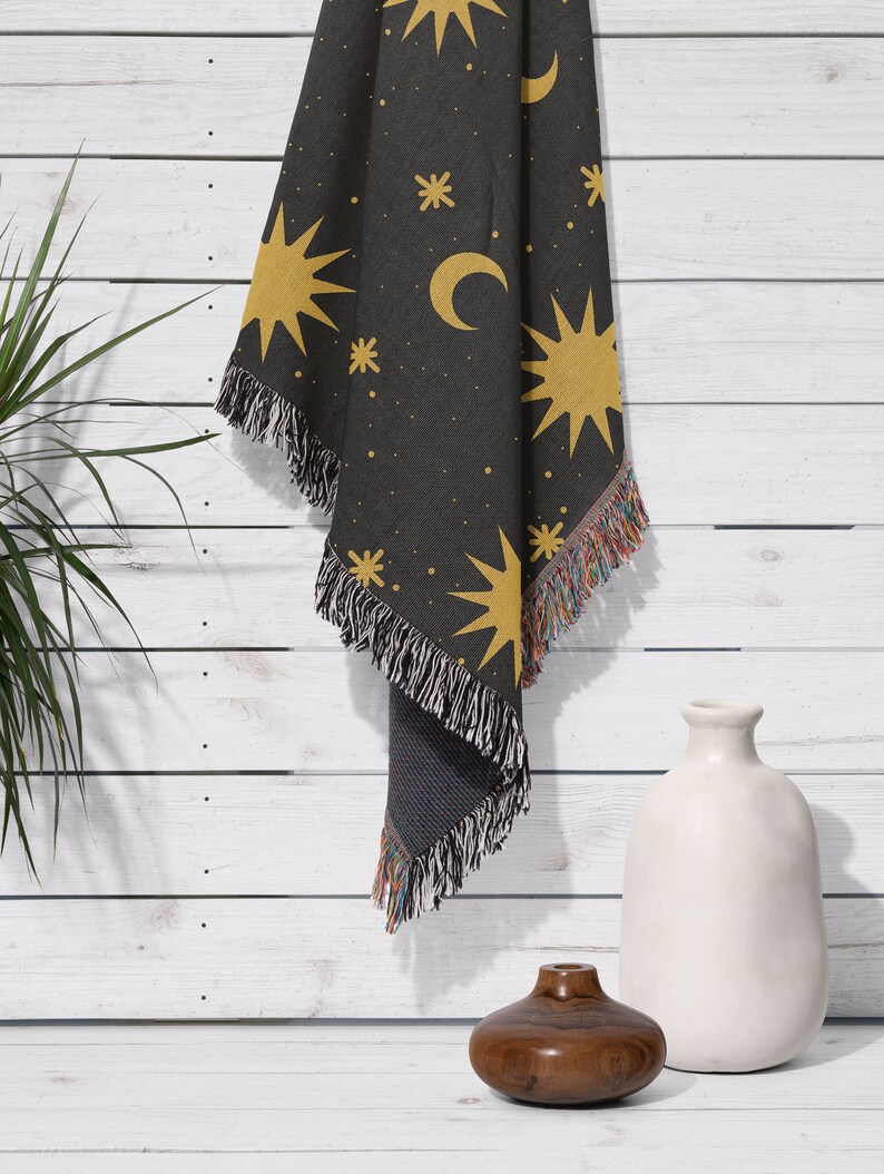 K&ouml;nnte beinhalten: Eine schwarze gewebte Wolldecke mit einem Muster aus gelben Sternen und Mondsicheln. Die Decke h&auml;ngt an einer wei&szlig;en Holzwand.