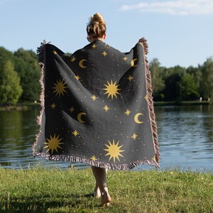 K&ouml;nnte beinhalten: Eine schwarze gewebte Wolldecke mit einem himmlischen Muster aus goldenen Sonnen, Monden und Sternen. Die Decke ist &uuml;ber die Schultern einer Person gelegt.