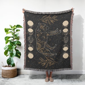 May include: A black woven throw blanket with a gold wolf design and moon phases. The wolf is surrounded by stars and a gold leaf design.