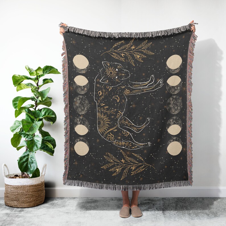May include: A black woven throw blanket with a gold wolf design and moon phases. The wolf is surrounded by stars and a gold leaf design.