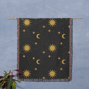 K&ouml;nnte beinhalten: Eine schwarze gewebte Wandbehang mit einem sich wiederholenden Muster aus goldenen Sternen und Mondsicheln. Der Wandbehang h&auml;ngt an einer hellblauen Wand.