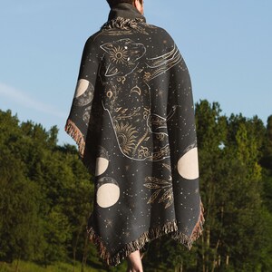 May include: A person wearing a black woven throw blanket with a gold celestial design featuring a wolf, moons, and stars. The blanket has fringe on the bottom.