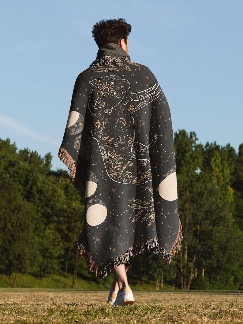 May include: A person wearing a black woven throw blanket with a gold celestial design featuring a wolf, moons, and stars. The blanket has fringe on the bottom.