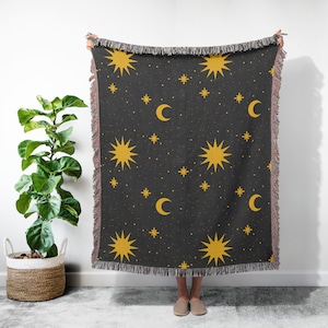 K&ouml;nnte beinhalten: Eine schwarze gewebte Wolldecke mit einem sich wiederholenden Muster aus gelben Sternen und Mondsicheln. Die Decke hat an allen Seiten Fransen.
