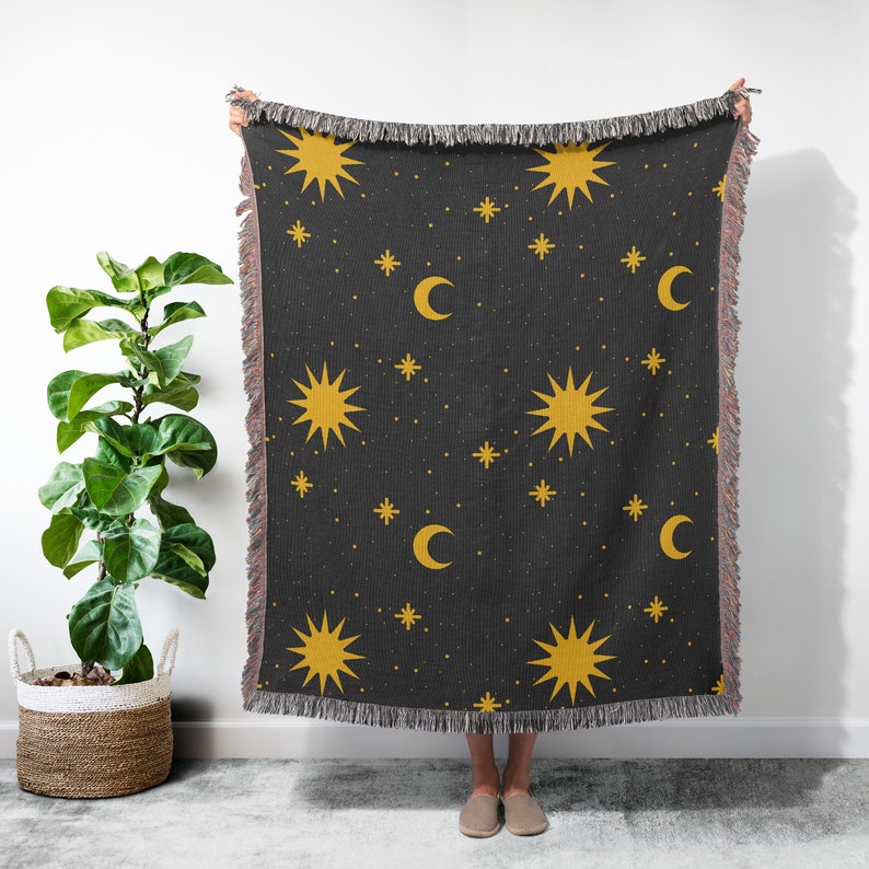 K&ouml;nnte beinhalten: Eine schwarze gewebte Wolldecke mit einem sich wiederholenden Muster aus gelben Sternen und Mondsicheln. Die Decke hat an allen Seiten Fransen.