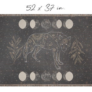 May include: A woven throw blanket with a dark gray background and a white wolf design. The wolf is surrounded by gold stars, moons, and leaves. The blanket measures 52 x 37 inches.