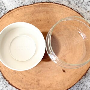 May include: Overhead shot of two bowls on a wooden surface. One bowl is white and opaque, while the other is clear glass. The bowls are placed on a round, light brown wooden slice, set against a speckled gray background.