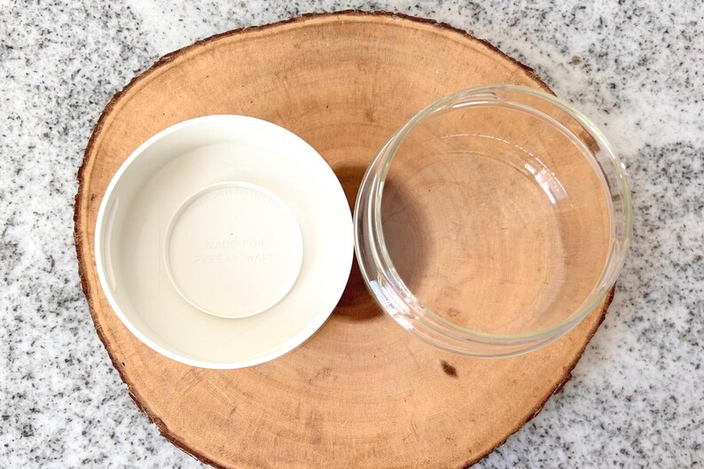 May include: Overhead shot of two bowls on a wooden surface. One bowl is white and opaque, while the other is clear glass. The bowls are placed on a round, light brown wooden slice, set against a speckled gray background.