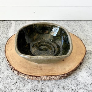 May include: A small, square ceramic bowl with a dark green and black glaze. The bowl is sitting on a wooden base.