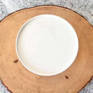 May include: A white ceramic plate on a round, light brown wooden serving board. The plate is centered on the board, creating a simple, natural aesthetic. The wood grain is visible, adding texture to the composition.