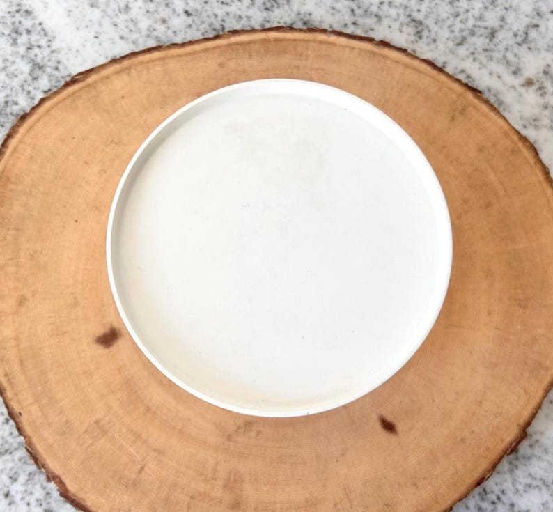 May include: A white ceramic plate on a round, light brown wooden serving board. The plate is centered on the board, creating a simple, natural aesthetic. The wood grain is visible, adding texture to the composition.