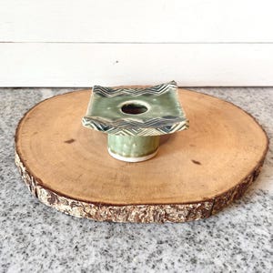 May include: A handcrafted ceramic ikebana flower frog in shades of green and white, with a square top and a circular opening. The top features a zigzag pattern. It sits on a round wooden slice.