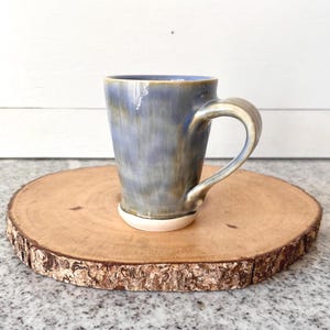 May include: A handmade ceramic mug with a blue and tan glaze, featuring a curved handle. The mug sits on a round, natural wood slice with a bark edge. The mug's base is white.