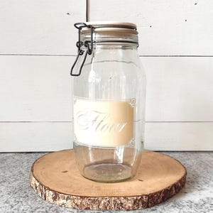 May include: A clear glass jar with a metal clasp and a wooden lid. The jar has a label that reads "Flour" in an elegant script. The jar sits on a round wooden slice, perfect for kitchen storage.