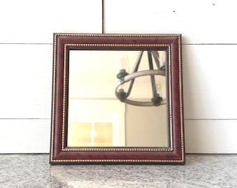May include: Square mirror with a burgundy frame and gold beaded trim. The mirror reflects a chandelier and a light-colored wall. The frame has a textured appearance, adding a touch of elegance to the design.
