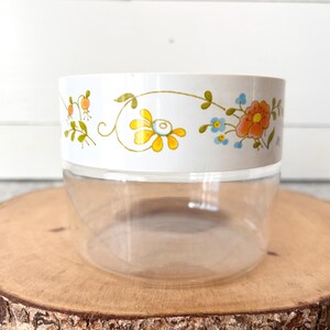 May include: A clear, round container with a white band featuring a floral design in yellow, orange, and blue. The container sits on a wooden slice, offering a rustic contrast to the vintage-style jar.