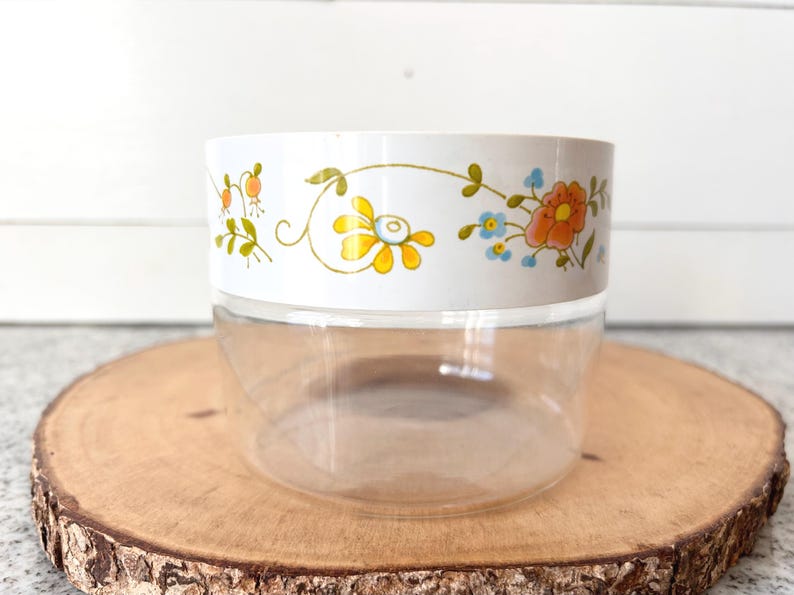 May include: A clear, round container with a white band featuring a floral design in yellow, orange, and blue. The container sits on a wooden slice, offering a rustic contrast to the vintage-style jar.