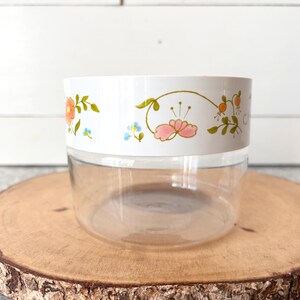 May include: A clear glass jar with a white lid featuring a floral design in orange, pink, and blue. The jar sits on a round wooden slice, showcasing a vintage aesthetic. The floral pattern adds a touch of color and charm to the container.