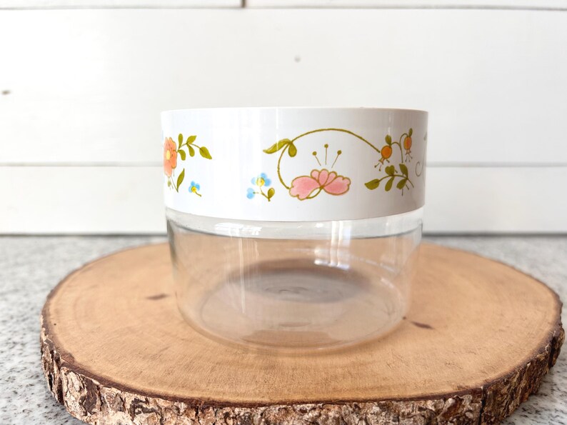 May include: A clear glass jar with a white lid featuring a floral design in orange, pink, and blue. The jar sits on a round wooden slice, showcasing a vintage aesthetic. The floral pattern adds a touch of color and charm to the container.