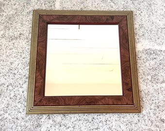 May include: Square mirror with a brown wood grain frame and gold trim. The mirror's frame has a rich, dark brown wood grain pattern, complemented by a thin gold border. The mirror itself reflects a soft, off-white hue.