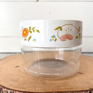 May include: A clear glass jar with a white lid featuring a floral design in orange, pink, and blue. The jar is sitting on a wooden slice. The text on the jar reads "MADE IN USA not for freezer or range top use."