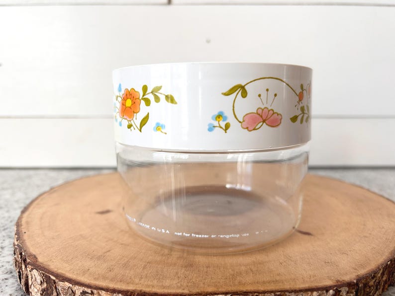 May include: A clear glass jar with a white lid featuring a floral design in orange, pink, and blue. The jar is sitting on a wooden slice. The text on the jar reads "MADE IN USA not for freezer or range top use."