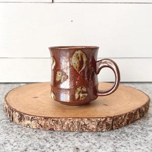 May include: A handmade ceramic mug with a brown glaze and leaf-like patterns in cream and tan. The mug has a rounded handle and sits on a wooden slice, perfect for enjoying coffee or tea.