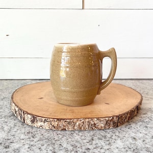 May include: A tan ceramic mug with a handle, sitting on a round wooden slab. The mug has a textured surface and a slightly flared rim. The wooden slab has a natural bark edge and is placed on a speckled surface.