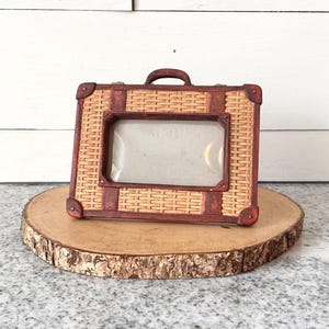 May include: A rectangular photo frame with a woven wicker design and a dark brown border. The frame has a handle and is displayed on a round wooden slice. The frame's corners are reinforced with dark brown accents.