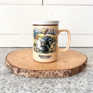 May include: Tan ceramic mug with a handle, featuring a colorful illustration of two bears, a bird, and trees. The mug has the text "Pigeon Forge Tennessee" on it. The mug is sitting on a wooden slice.