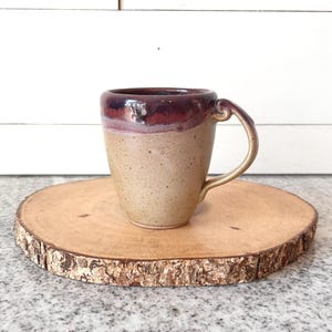 May include: A handmade ceramic mug with a burgundy rim and handle, and a speckled tan body. The mug is set on a round wooden coaster with a natural bark edge. The mug is designed for hot beverages.